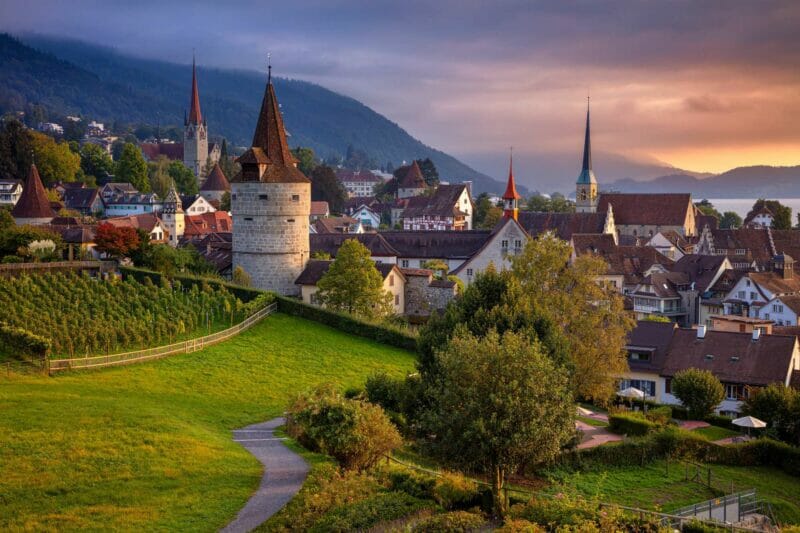 A pretty town with towers and red roofs and mountains in the background -  Zug