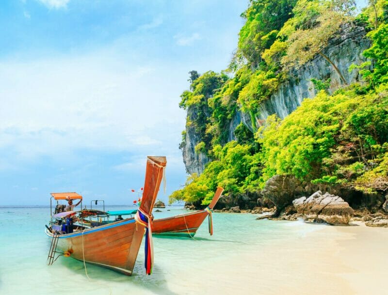 A tropical beach in Phuket - emerelad waters, colorful boats and a golden beach framed by rocky hills.