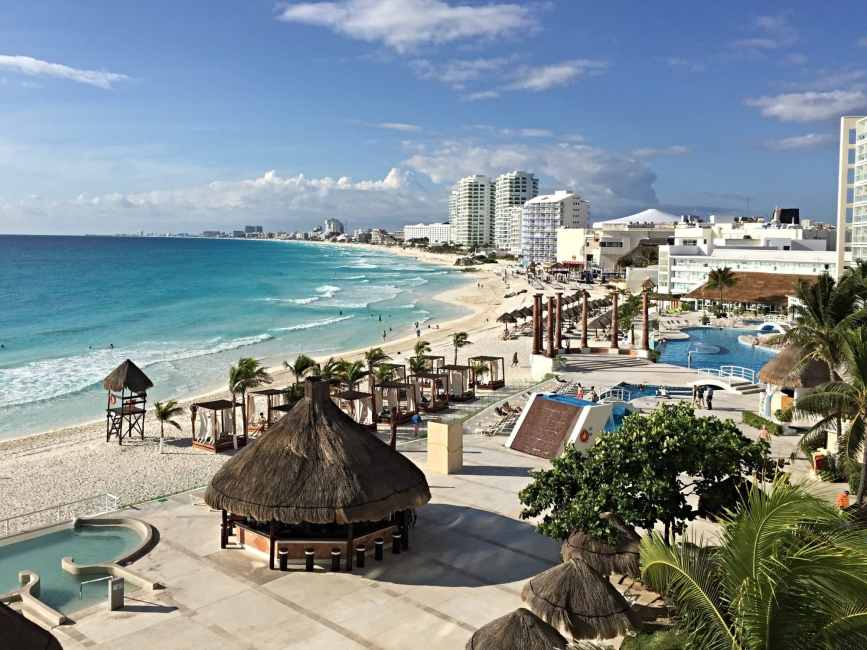 A sandy beach in Cancun with apartment blocks and hotels with palm trees and pools along the beach line